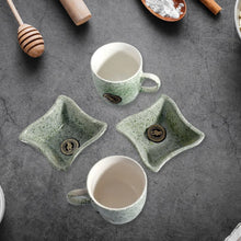 Two green ceramic mugs and two matching small bowls on a gray surface with kitchen items around.