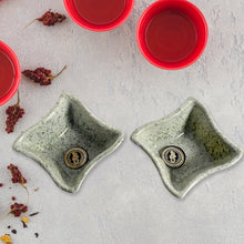 Two green ceramic dishes with a logo on a light gray surface, surrounded by red cups and dried red peppers.