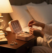 Person organizing jewelry in a pink jewelry box on a bedside table with warm lighting.