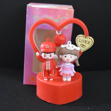 Figurine of a boy and girl holding a heart with a 'Love' card, standing on a red base against a dark background.